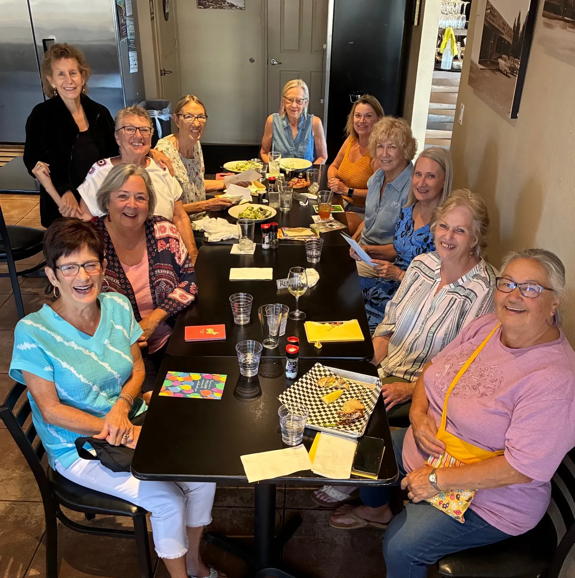 A group of eleven older women sit around a restaurant table with food, drinks, and gift cards, smiling at the camera.