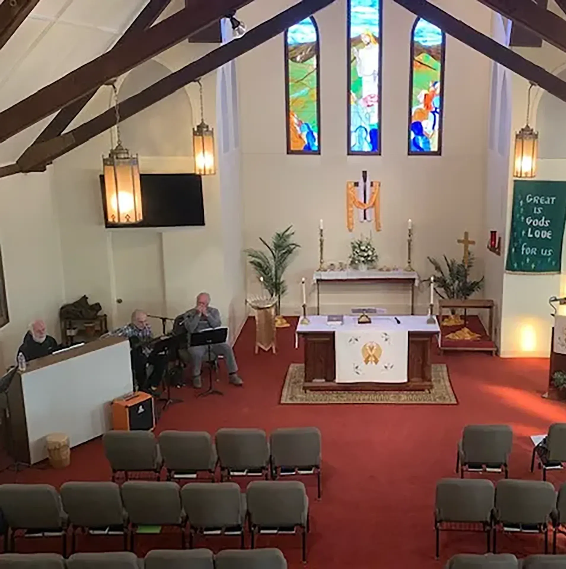 Interior of a small church with rows of chairs, altar at the front, musicians to the left, stained glass windows, and a banner reading,