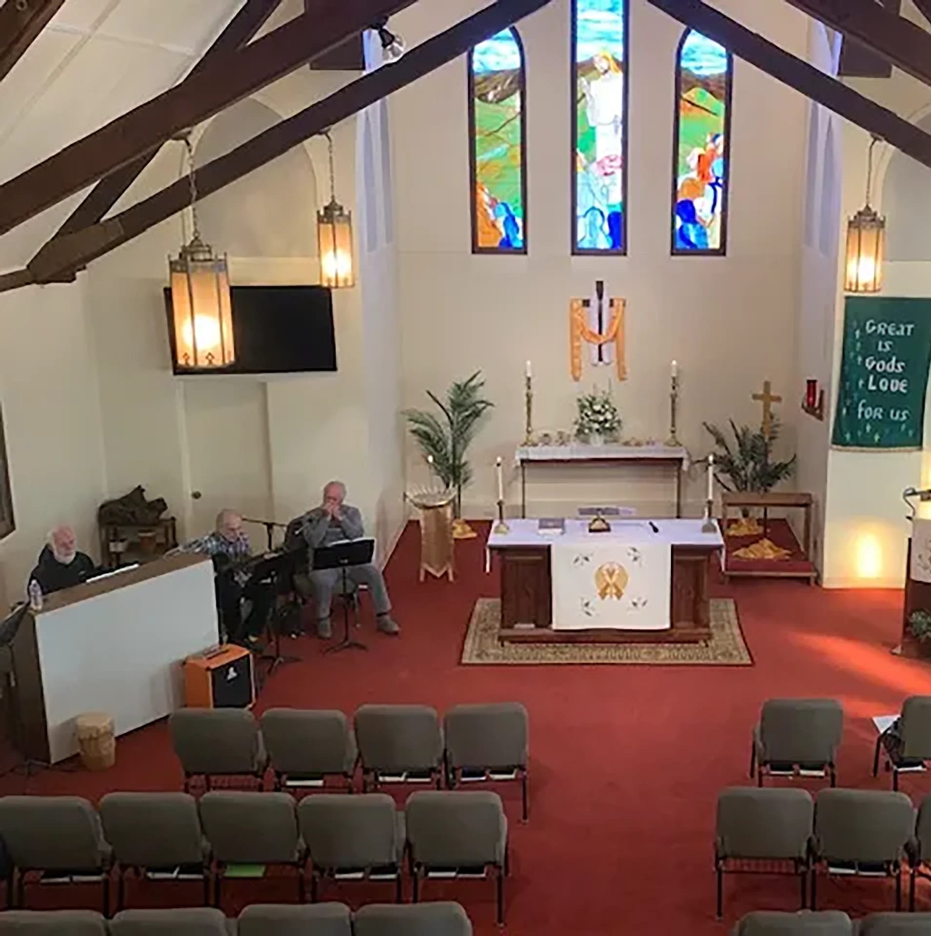 Interior of a small church with rows of chairs, altar at the front, musicians to the left, stained glass windows, and a banner reading, "Great is God's love for us.